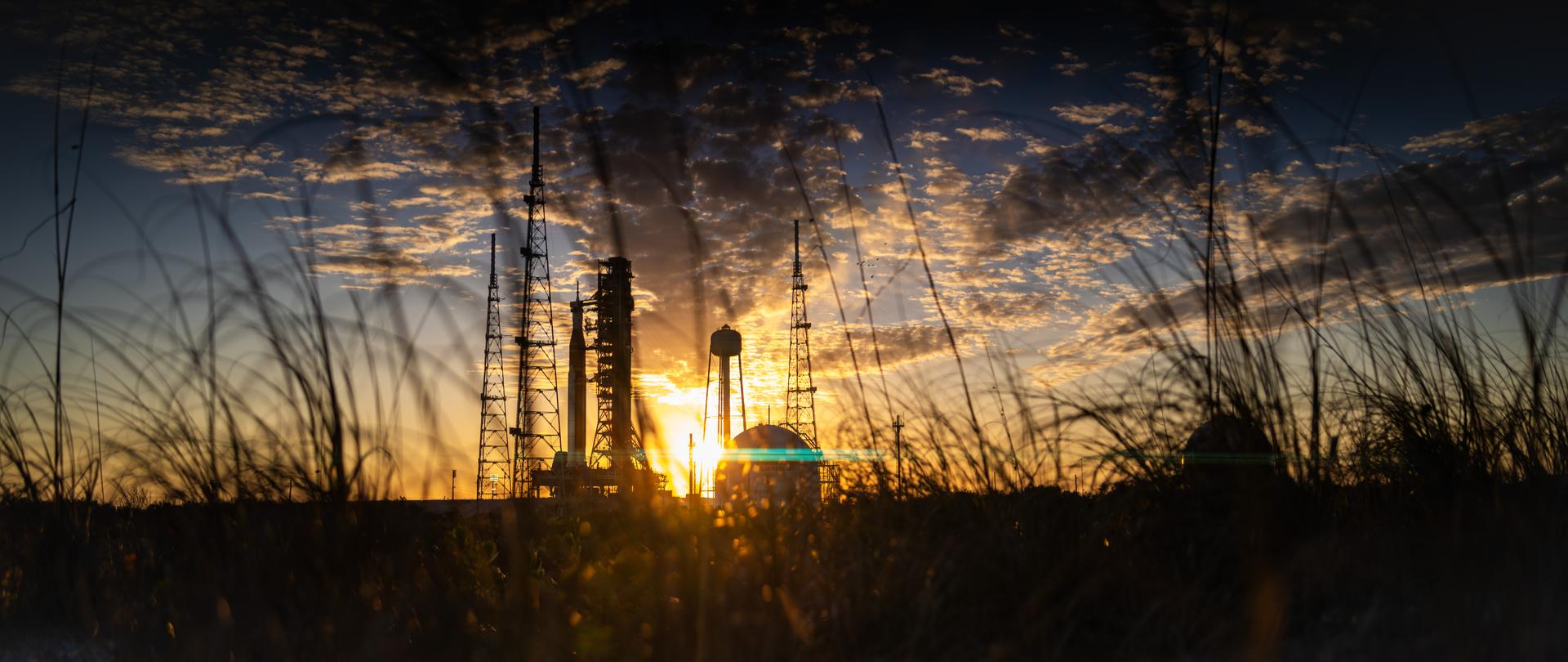 The sun sets behind NASA’s Space Launch System (SLS) rocket and Orion spacecraft as they stand fully assembled atop the mobile launcher at Launch Pad 39B at NASA’s Kennedy Space Center in Florida. The sky glows with warm shades of orange and pink, silhouetting the towering rocket and its solid rocket boosters against the fading light.  Photographed on January 31, 2026, the scene captures teams preparing for a wet dress rehearsal for the Artemis II mission, rehearsing launch countdown timelines and procedures as day turns to night. 